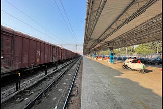 A railway station in the afternoon in a remote part of India. The image is divided into four quadrants with tracks and the platform on the bottom, the roof of the platform and a blue sky above, and everything meets at the vanishing point directly in the center.