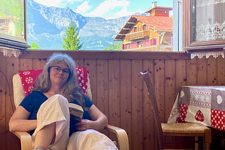 A woman sits on a chair with a book. Mountains and the top of a mountain chalet are visible through the window behind her.