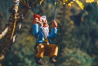 A gnome in a blue jacket and red hat sitting on a tree swing in the sunshine