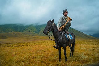 An indiginous man sitting on the back of a black horse in an open land filled with green grass with a mountain in the background and lots of clouds in the sky