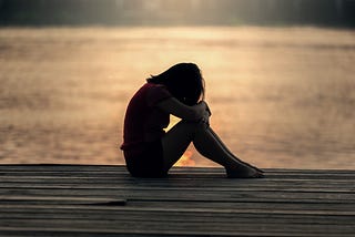 A young woman sitting on the pier with the sea behind her, curled up looking down feeling sad and ashamed