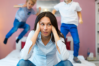 A woman holding her head; two children jumping on bed behind