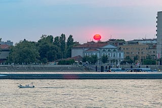 A bright red sun floats over historic buildings and a river. A boat floats on the river.