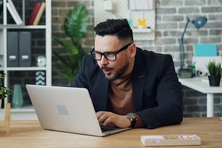A man in glasses and a blazer leans toward a laptop, appearing focused during a video call in a modern office. He is surrounded by shelves containing folders and a brick wall is visible in the background.