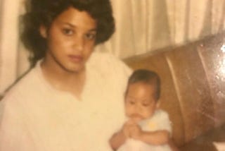 A sepia toned photo of a beautiful Black woman with light brown skin and voluminous black hair holding a baby.