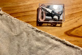 A piece of unevenly stitched linen on a wooden surface next to a small clear rectangular sewing kit full of sewing supplies like scissors, thread, and measuring tape.