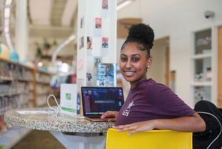 Teen sitting in library with laptop, smiling at the camera