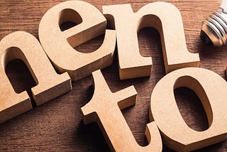 Wood block letters are arranged on a table next to a lightbulb spelling the word “mentor” on a dark wood table.