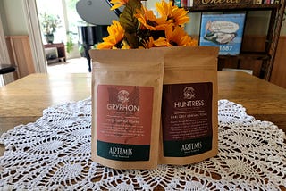 Two tan kraft paper bags of tea sitting on a doily in the middle of an oak table, with a bouquet of yellow sunflowers behind them, and a glossy black grand piano in the background.