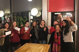 Photo of 8 women smiling, some holding up cards, some waving at camera. In a cafe, standing up behind table.