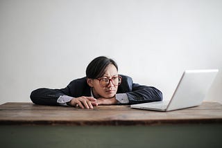 A man dressed in a blue suit wearing red glasses with his chin on his wrists, staring defeatedly at an open laptop.