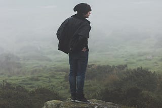 Photo of a man in a black hat and black coat and jeans standing on a ledge looking out over a foggy field