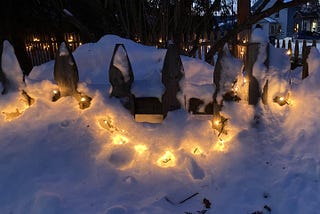 Fairy lights shining through snow outdoors hanging from a white picket fance