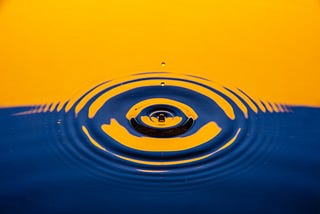 A close up of a water droplet piercing the stillness of a river reflecting orange and blue back hues.