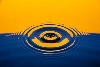 A close up of a water droplet piercing the stillness of a river reflecting orange and blue back hues.