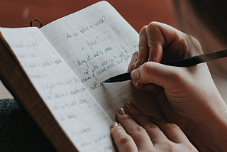 Picture is close in of woman’s hands holding and writing with pen in journal.