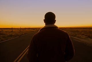 A young man stands in the middle of a deserted highway, looking away from the camera down the road. The horizon glows with the orange light of dawn.