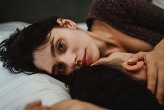 Couple in bed together, the female partner staring straight into the lens of the camera