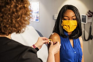 a Black woman wearing a blue sleeveless top receives a bandaid after receiving a vaccination from a white-appearing woman whose face is away from the camera