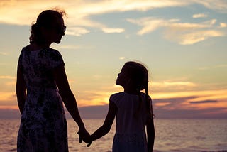 A young mother holds hands of her young daughter on a beach.