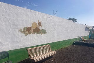 There is a white wall running diagonally away on which is painted a lying down lurcher or greyhound type dog. Below, where a green stripe running beneath it meets the white paint, is a bench, and over the top of the wall, in the distance, is the top of a pine tree. This is Sara.
