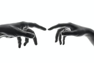 Two people’s hands face one another, a larger left hand on the left and a smaller right hand on the right; the pointer fingers are extended and nearly touching.