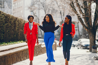 Three women of color are walking on a sidewalk. On the far left is a woman in a red suit with her hair pulled back, center is a woman in a black blouse and cobalt blue pants gesturing with her hands, on the right is a woman dressed in all black with a red blazer; her hair is braided and pulled back.