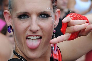 punk person with purple earrings and heavy eye makeup, sticking out their tongue and pointing to it