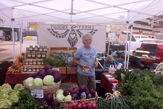 A woman farmer with gray hair at a booth full of vegetables at a Farm Stand.