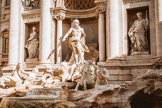 A view of the Trevi Fountain. Sculptures of roman gods and horses sculpted in white marble are touched by gentle ray of sun.