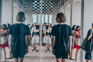 A person in black t-shirts stands facing a large mirrored wall in a modern architectural space with concrete beams and checkered ceiling panels. His reflections are visible in the mirror, along with a crowd of visitors in the background. The setting appears to be a contemporary museum or cultural institution with industrial design elements and tiled flooring.