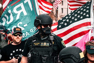 A policeman wearing riot gear in front of a men holding American and Trump flags.