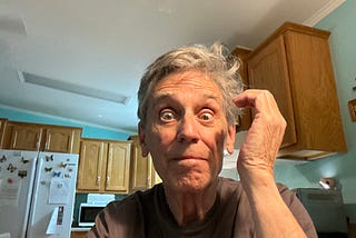 An older man with gray hair and a surprised, wide-eyed expression is sitting in a kitchen. He is wearing a dark t-shirt and has one hand raised to his head, as if scratching or emphasizing confusion.