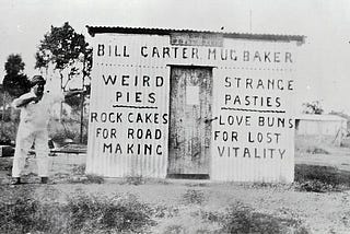 An old photograph of a man standing next to the side of a shed, on which is painted the words: “Bill Carter Mug Baker”