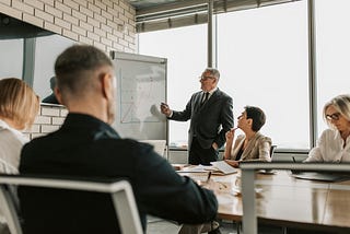 A man in a business suit at a whiteboard presenting something, while the rest of the team in business formal attire watches.