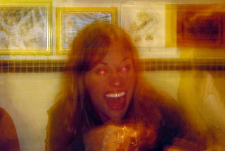 Uma mulher com a boca aberta no centro com os olhos rosados-vermelhos meio desfocado como uma foto tirada em uma longa exposição onde há movimento.