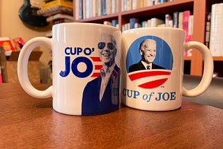 Two coffee mugs sitting on a dining table, one featuring a picture of Joe Biden inserted into the center of the Obama 2012 campaign logo with the text “Cup of Joe” written underneath, the other featuring a smiling portrait of Biden in his signature sunglasses, with the words “Cup o’ Joe” next to it.