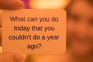 A closeup of someone holding a small sign saying ‘What can you do today that you couldn’t do a year ago?’.