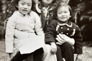 Smiling young woman kneeing down with two smiling girls, one in each arm.