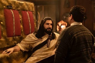 Nandor The Relentless (Kayvan Novak) in a fur-lined coffin with the lid propped open, slightly sitting up and talking to his Familiar, Guillermo (Harvey Guillen).