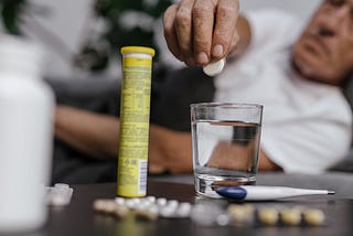 Someone is about to drop a tablet into a glass of water. There is a thermometer and medication on the table near them