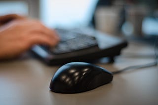 A computer mouse on a desk with hands on a keyboard slightly blurred in the background.