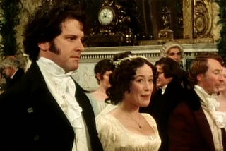 Uma cena da adaptação de Orgulho e Preconceito de 1995, estrelando Jennifer Ehle como Elizabeth Bennet e Colin Firth como Sr. Darcy. Podemos ver o Sr. Darcy e Elizabeth Bennet dançando em um baile, com várias pessoas acompanhando a dança ao fundo. O Sr. Darcy tem a “cara fechada”, como se estivesse desconfortável, enquanto Elizabeth parece sorrir com desdém enquanto lhe diz algo.