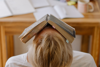 Overwhelmed student surrounded by books sleeping with book on head