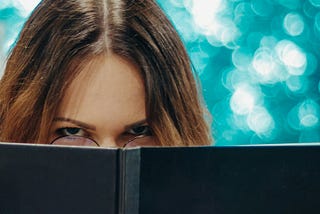 The image features a woman peeking over the top of an open book, with a mysterious expression and glasses slightly visible. The background is a soft blur of teal and white, creating a dreamy and reflective atmosphere.