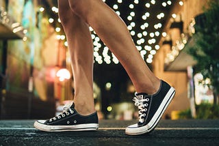 Two thin legs from the knees down, mid-walking stride, in cute black converse low-top sneakers and a lovely street in the background