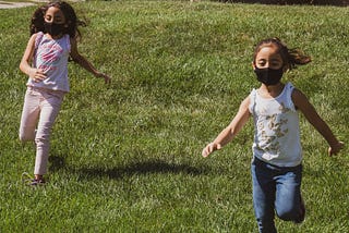 two girls wearing cloth face masks and playing on a green grass field during daytime