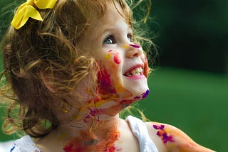 A two or three year old girl smiling at her parent or caregiver with paints on her face and dress.