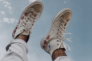 A person’s cuffed jeans and white converse with red heart designs on them raised toward a light blue sky with wispy clouds