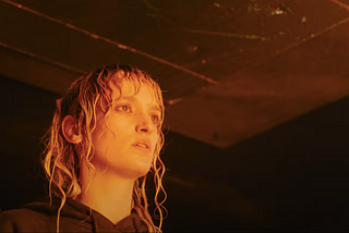 The protagonist of Titane, Alexia with soaked hair in a black hoodie, looking off into the distance in a garage, awash in an orange glow.
